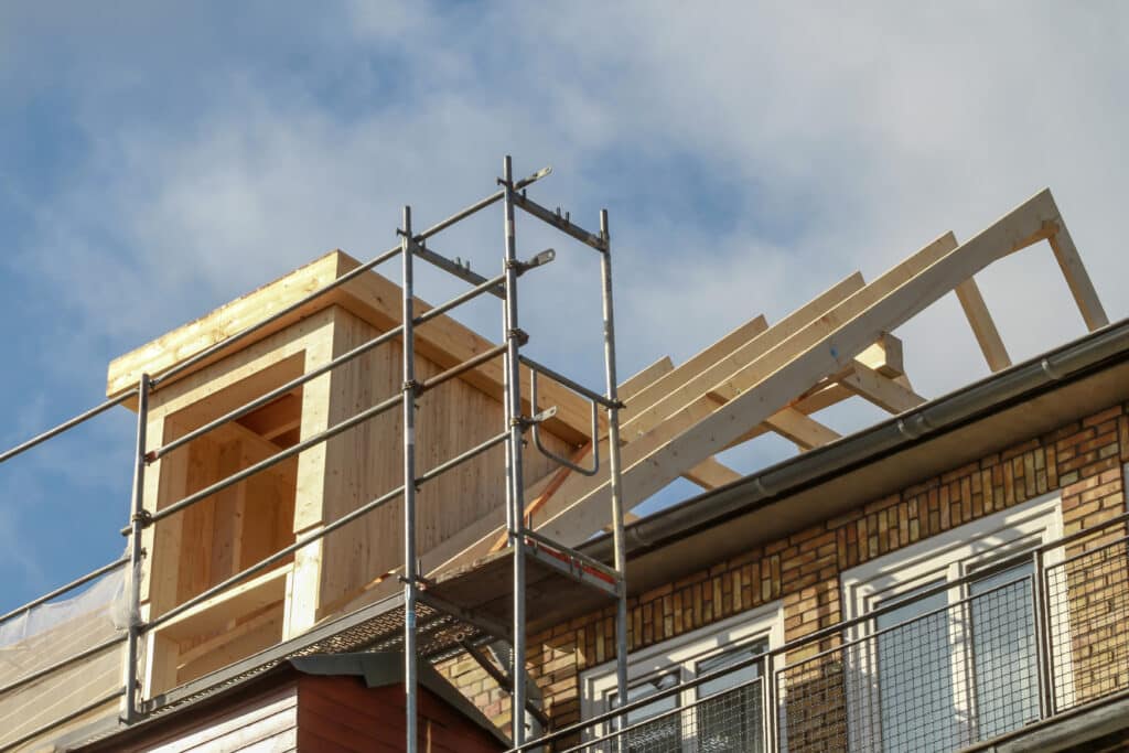 Construction toit: Lucarne en bois, charpente et rénovation Construction de lucarne et charpente en bois sur un toit d'immeuble, avec échafaudage, sous un ciel bleu nuageux.