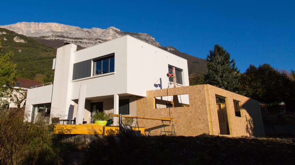 Maison moderne en construction, vue sur montagne Maison moderne blanche en construction avec une extension en bois, sous un ciel bleu. Une grande montagne rocheuse domine l'arrière-plan.