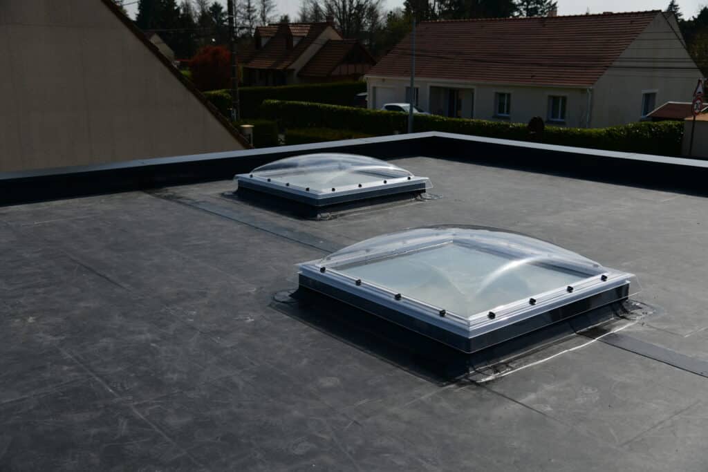 Toit plat avec Velux : Éclairage naturel et étanchéité. Toit plat étanche noir avec deux puits de lumière Velux installés. En arrière-plan, maisons résidentielles et panneau 30 km/h.